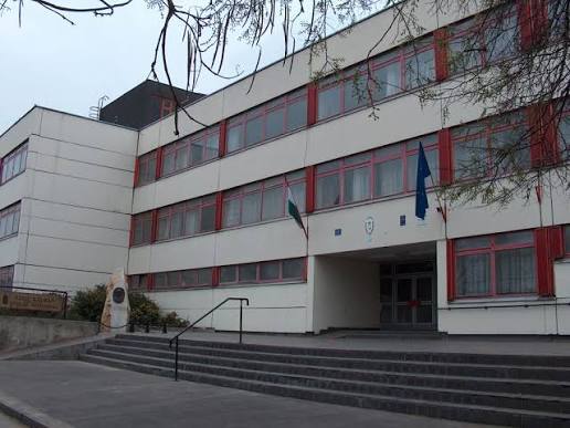 A Szily Kálmán Technikum főbejárata, piros ablakkeretes homlokzat, lépcsősor és zászlók.